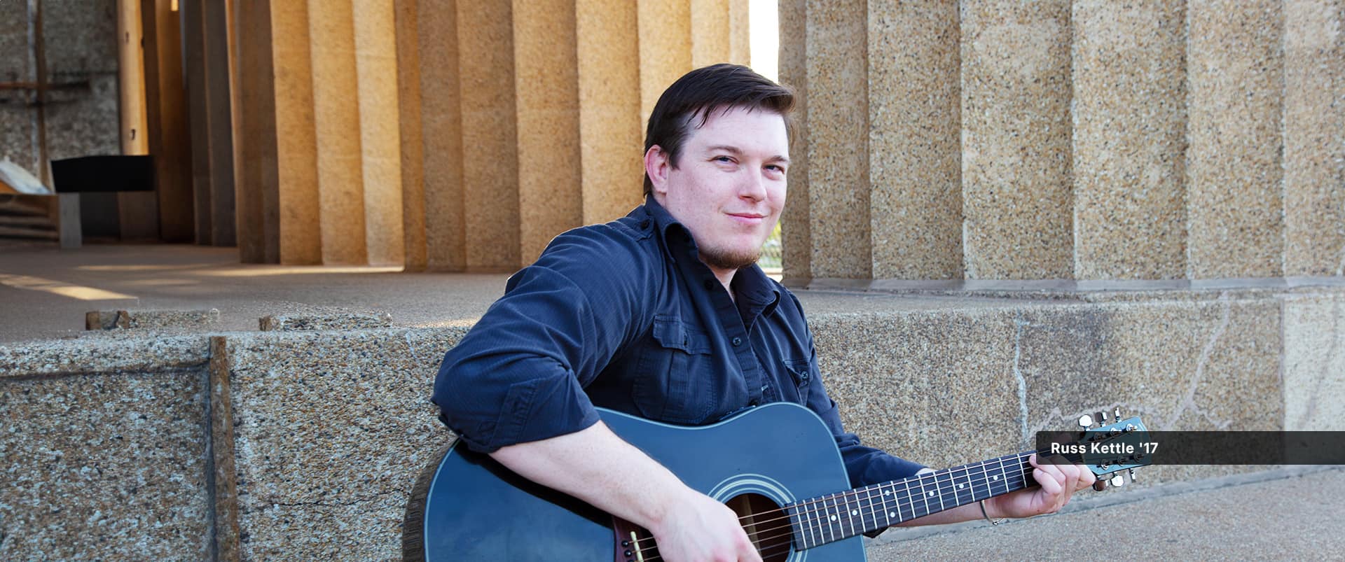 Russ Kettle, who earned his degree in 2017, wearing an unbuttoned button-down shirt and black T-shirt holding a guitar.