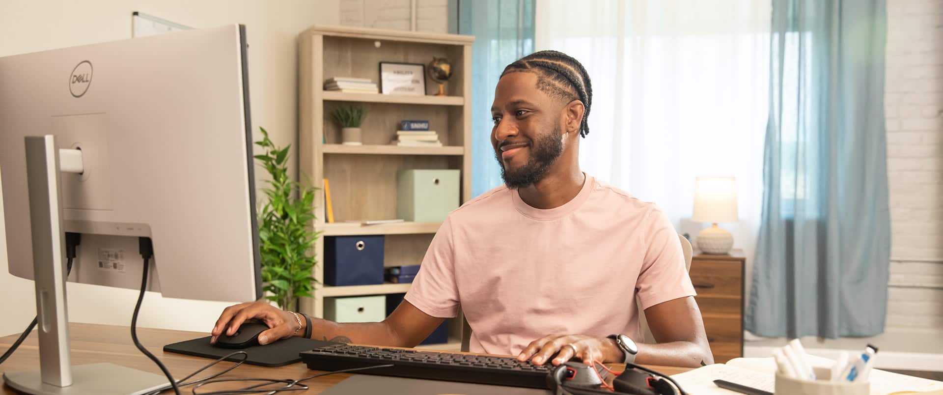 Matthew Seawright, who earned his degree in 2019, using a desktop computer in a small office with a bookcase, lamp and floor plant in the background