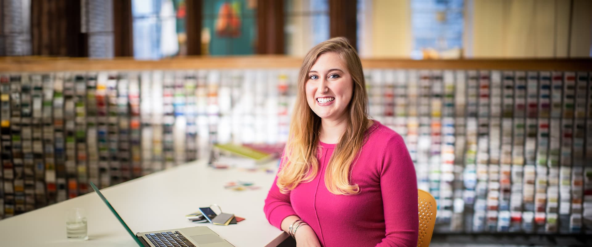 Caitlyn Carey, who earned her degree from SNHU, wearing a bright pink sweater standing at a table with an open laptop beside her.
