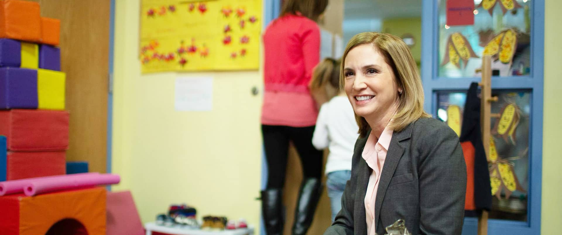 Marti Ilg, who earned her degree from SNHU 2014, sitting in a elementary school classroom with children's art on the wall and a woman leading a child out of the room by the hand in the background.