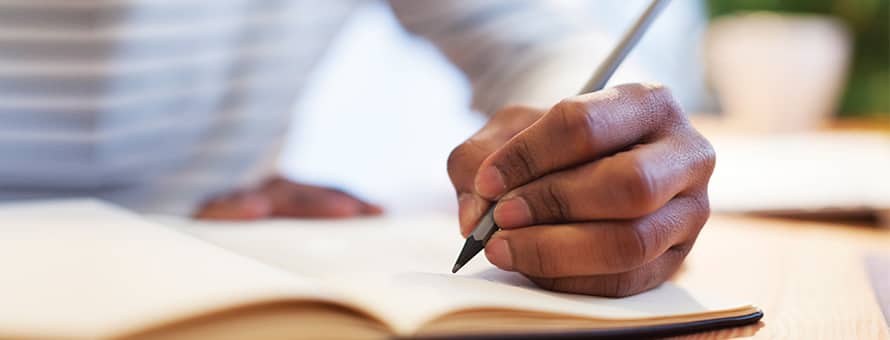 The hand of an MFA student holding a pencil and writing in a notebook