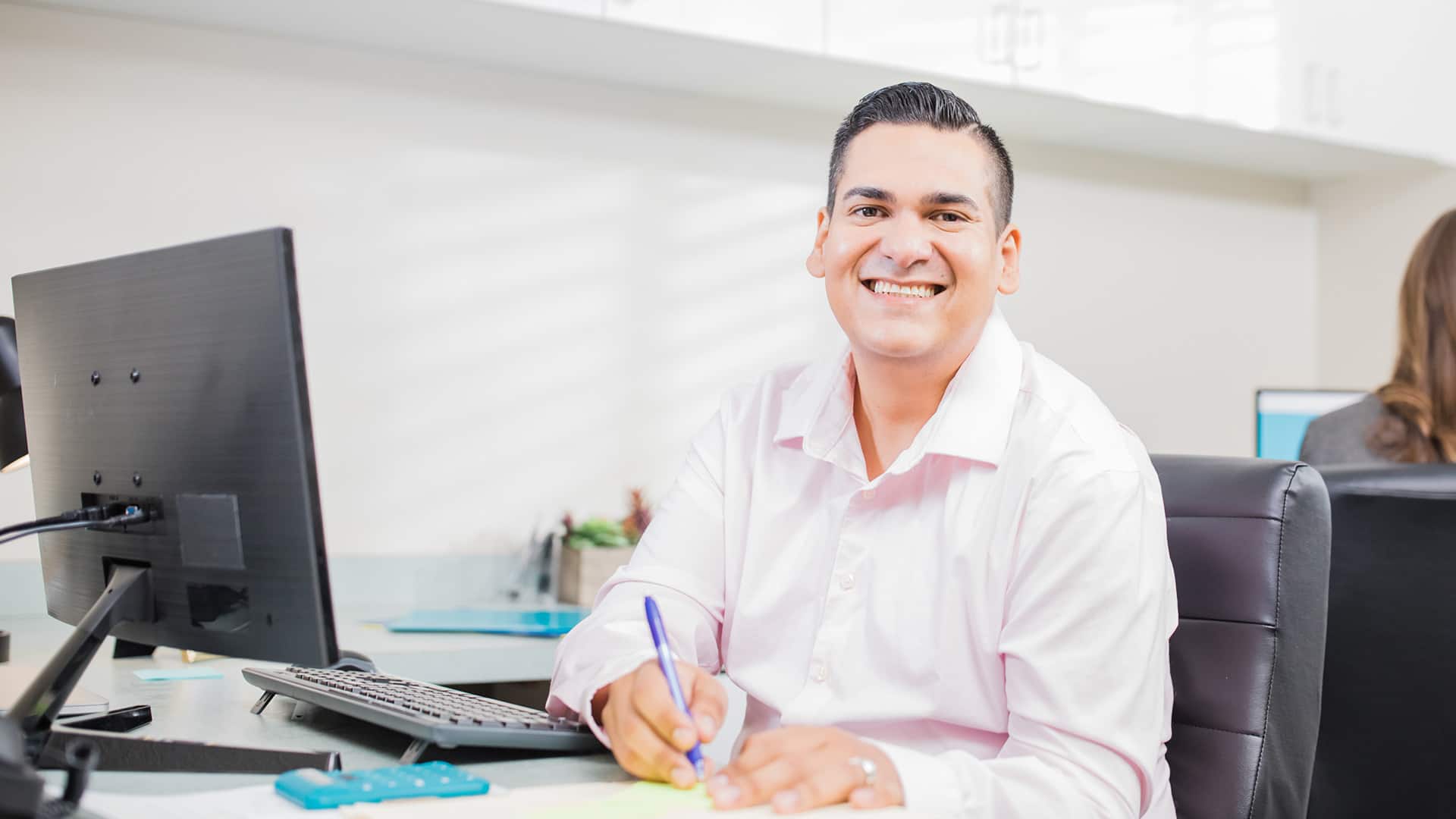 Juan Munoz, who earned his MBA from SNHU in 2023, wearing a button down shirt sitting at a desk with a computer monitor and holding a pen.
