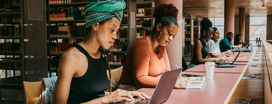 Two master's students studying in a library, one using a laptop and the other reading a textbook.