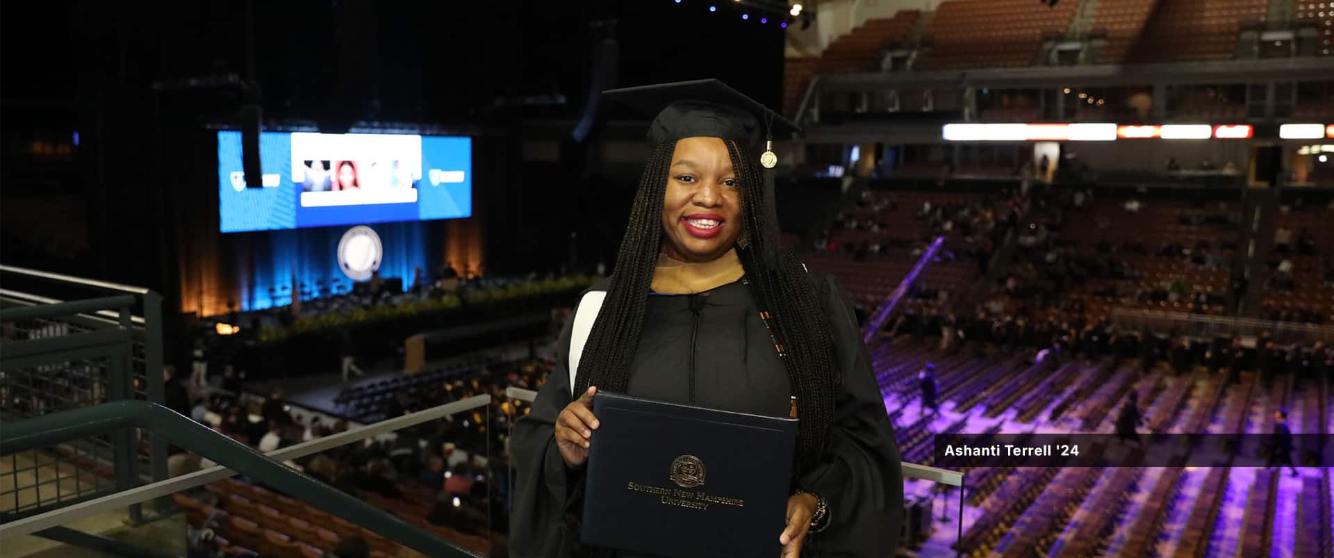 Ashanti Terrell, who earned her master's in IT from SNHU in 2024, wearing her cap and gown and holding her diploma on a balcony above the SNHU commencement floor.