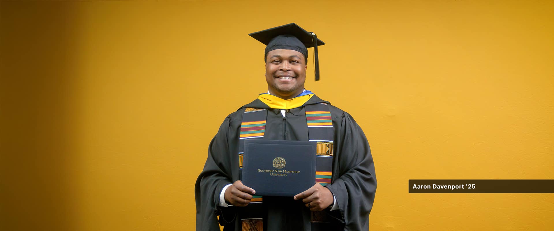 Aaron Davenport, who earned his master's in finance from SNHU in 2025, wearing his cap and gown and holding his diploma in front of a yellow backdrop.