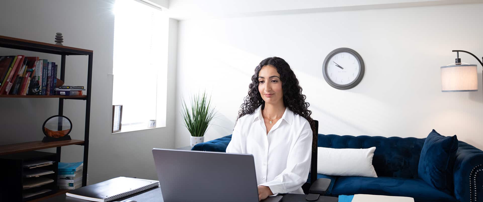 Fatma Salem Pease, who earned her degree from SNHU, wearing a white button down shirt sitting at an office desk typing on her laptop with a blue couch, wall clock and bookshelves in the background.