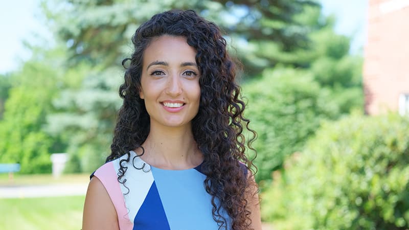 Fatima Salem Pease, who earned her degree from SNHU in 2019, standing outdoors wearing a blue, white and pink  dress with green shrubs and trees in the background.