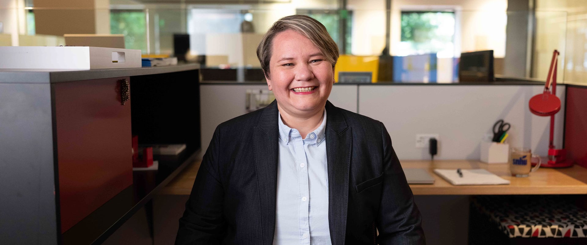Alaine Garcia, who earned her degree from SNHU in 2020, wearing a dark blazer and blue button down  shirt standing in an office cubicle.