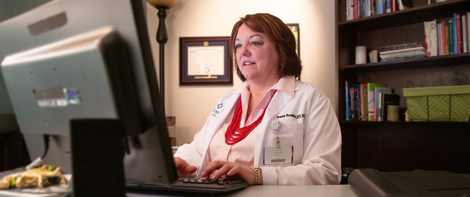 Bonnie Fecowicz, who earn her degree from SNHU, wearing a white lab coat, working on a desktop  computer at a desk with her framed SNHU diploma on the wall behind her.