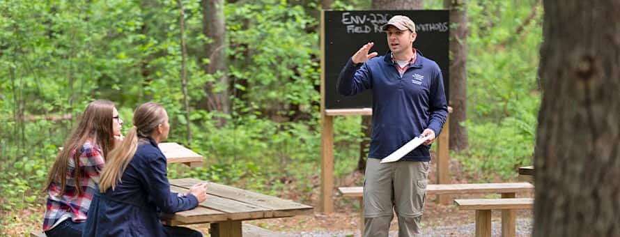 Arboretum Administrator Michael Weinstein holding a clipboard in the accredited arboretum, talking to two students at a picnic table.
