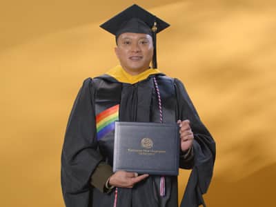 Prince Gelito, who earned his online master's in cybersecurity from SNHU in 2024, wearing his cap and gown and holding his diploma in front of a yellow backdrop.