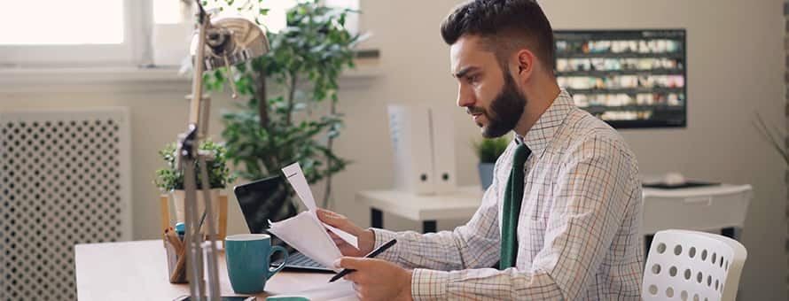 A person with a beard wearing a professional attire and examining paperwork at a desk.