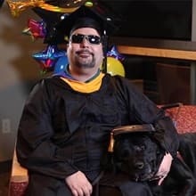 Ricardo Scarello, who earned his online master's in IT from SNHU in 2019, wearing his cap and gown with his guide dog beside him and balloons in the background.