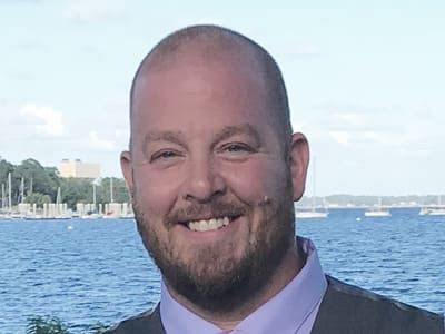 Rick McQueen, who earned degrees from Southern New Hampshire University in 2019 and 2021 standing in front of a body of water on a sunny day.