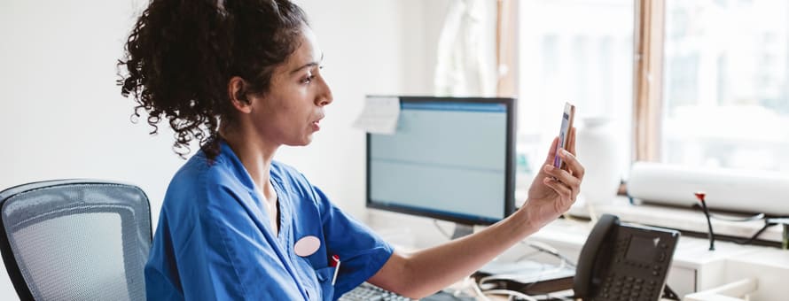 A woman with dark hair in a blue medical shirt looking at a phone and working in the role of telehealth in nursing