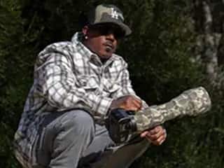 Ryan Carter, who took online photography and graphic design courses at SNHU, squatting in a wooded setting, holding a camera with a camouflaged lens.