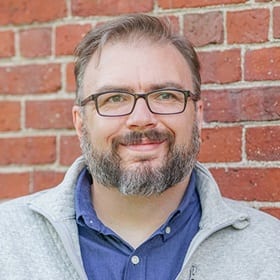 SNHU associate dean Seth Matthews in front of a red brick wall wearing a blue shirt, grey sweater.