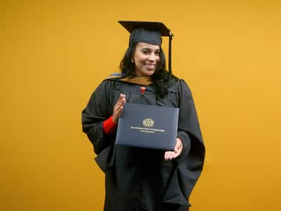 Sheralyn Hernandez, who eanred her online MBA in Healthcare Management from SNHU 2021, wearing her cap and gown and holding her diploma in front of a yellow backdrop.