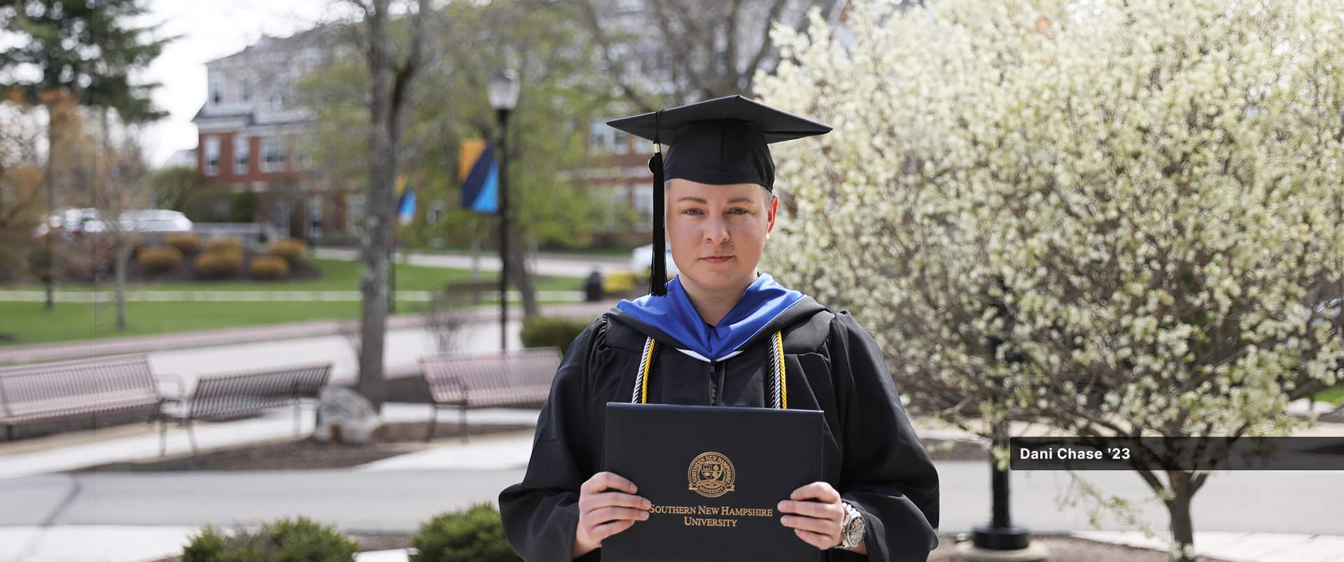 Dani Chase, who earned their bachelor's in sociology from SNHU in 2023, wearing their cap and gown and holding their diploma with benches and campus buildings visible in the background.