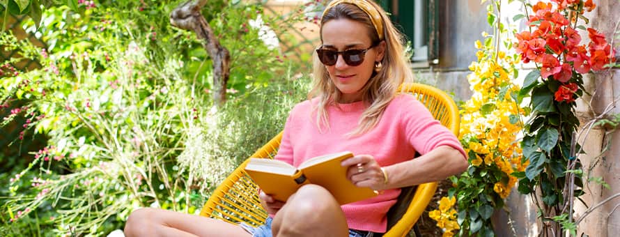 A woman with blonde hair in a pink shirt sitting in a yellow chair reading a book in the summer