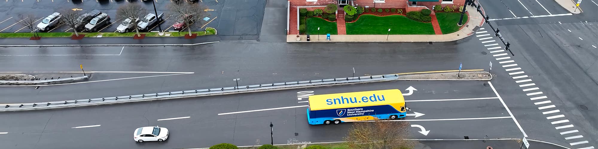 A yellow and blue coach bus with SNHU’s logo on the side seen from above at an intersection.