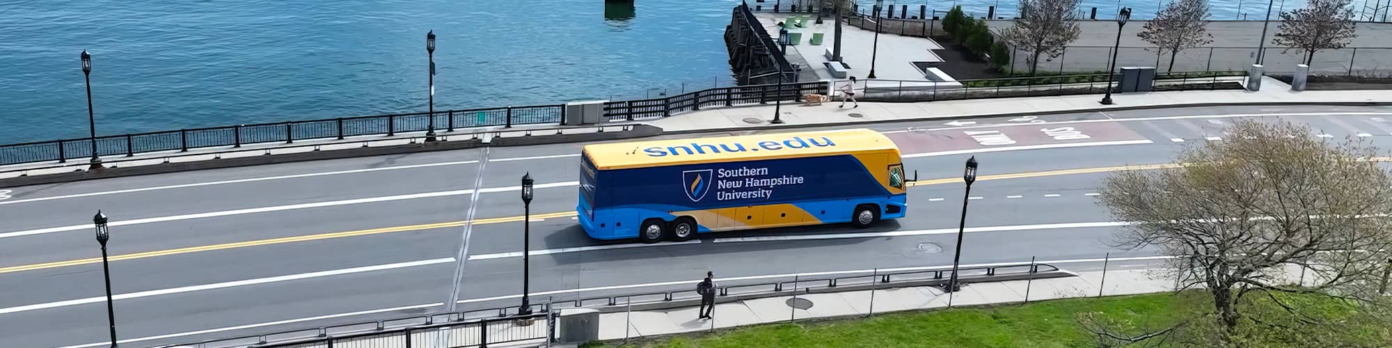 A yellow and blue coach bus with SNHU’s logo on the side seen from above driving over a bridge.