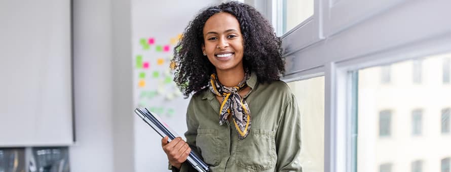A woman smiling while holding a laptop and notebooks in a modern office.