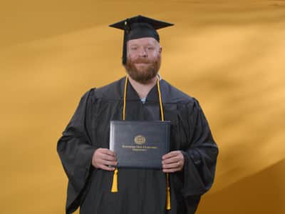 Travis Baril, who earned his online bachelor's in psychology from SNHU in 2024, wearing his cap and gown and holding his diploma in front of a yellow backdrop.