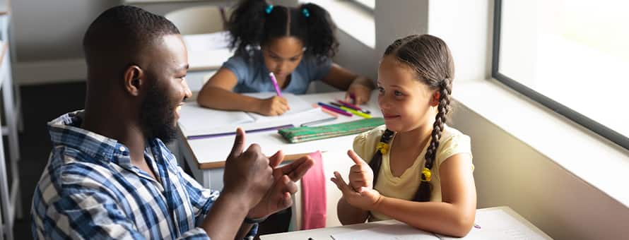 A male teacher kneeling and making eye contact while instructing a child in a classroom.