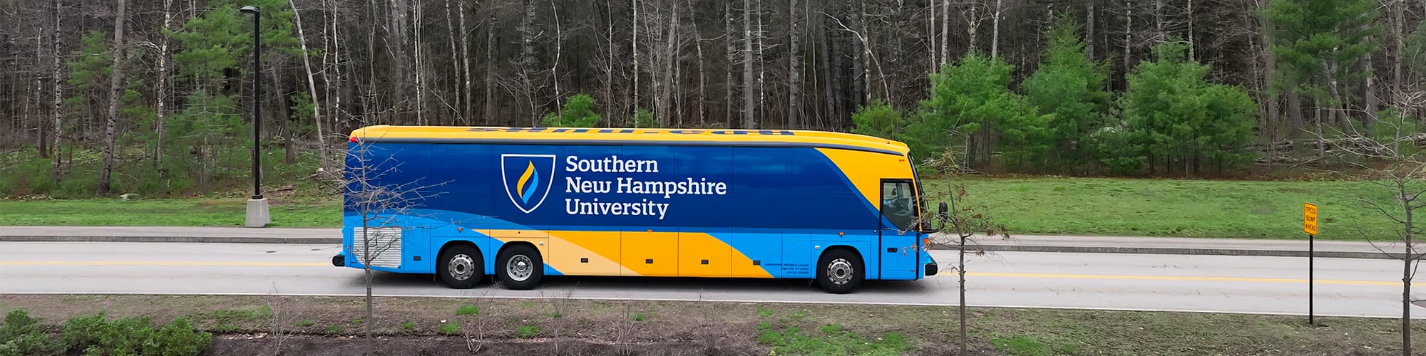 A yellow and blue coach bus with SNHU’s logo on the side driving on a small road with trees in the background.