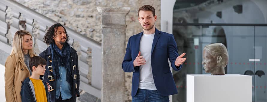 A curator with a master's in history, gesturing to a sculpture of a head during a museum tour.