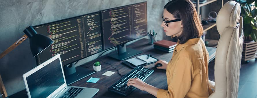 A cybersecurity analyst working at her desk with a laptop and two monitors with cyber code in front of her.