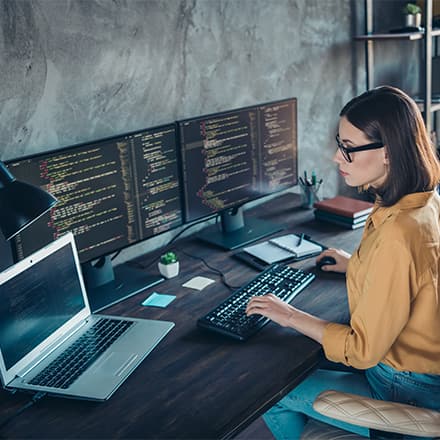 A cybersecurity analyst working at her desk with a laptop and two monitors with cyber code in front of her.