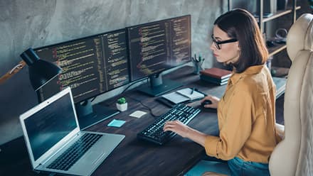 A cybersecurity analyst working at her desk with a laptop and two monitors with cyber code in front of her.