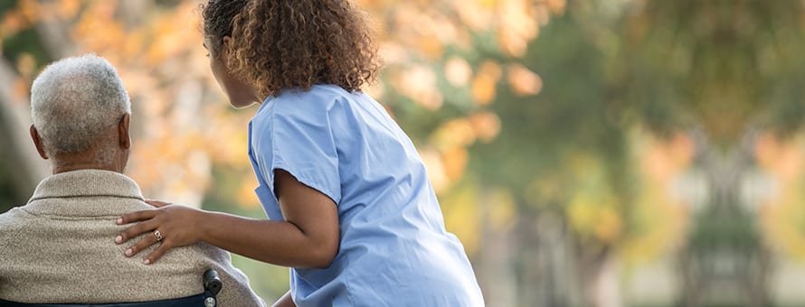 A CNA wearing blue scrubs with her hand on the shoulder of a patient in a wheelchair.