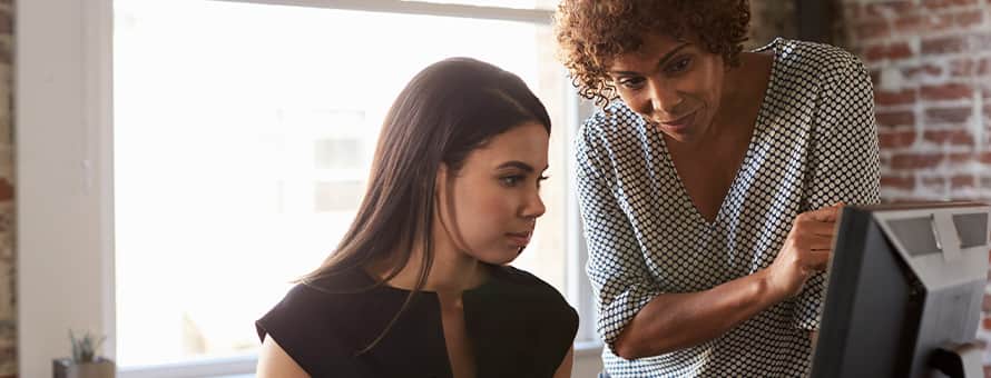 One woman sitting look at a desktop with another woman standing over her looking at the desktop