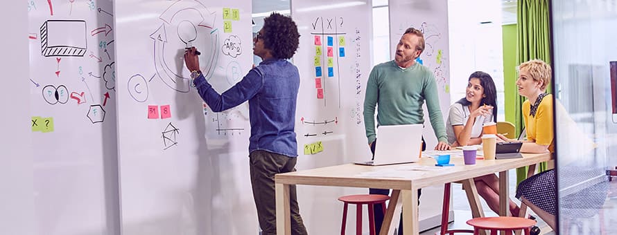 A group of students working towards their BA degree. One student is writing on the white board while three other students surrounding a desk watch. 