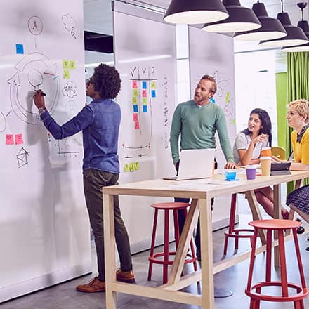A group of students working towards their BA degree. One student is writing on the white board while three other students surrounding a desk watch. 
