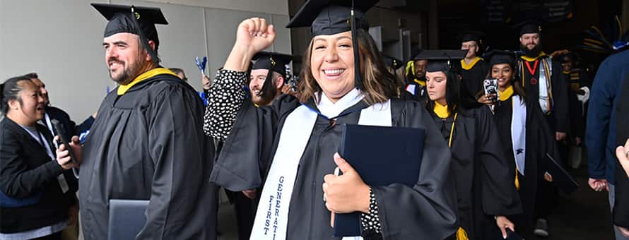 A graduate wearing a first generation stoll raising a fist and smiling while walking in a line of other graduates holding their degrees.