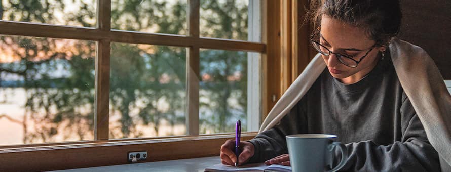 A speculative fiction writer with a mug, writing in a notebook beside a window overlooking a lake.