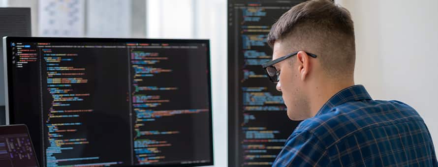 A computer science student with a laptop and two monitors of code in front of him, considering what master's degree to get.