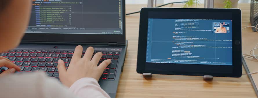 A person coding at a laptop while referring to a tablet featuring more code and a video call in the corner of the screen.